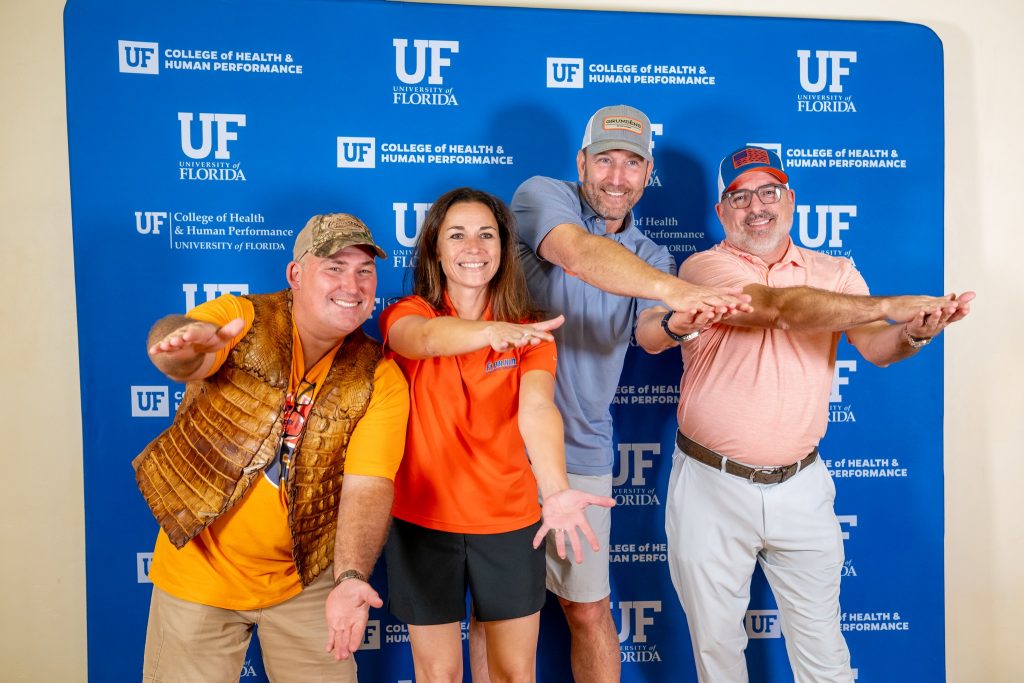 Four individuals dressed in Gator gear pose doing the Gator chomp in front of a UF College of Health & Human Performance step and repeat.