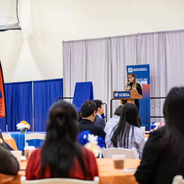 A presenter speaks at the podium in front of a large crowd seated at tables.